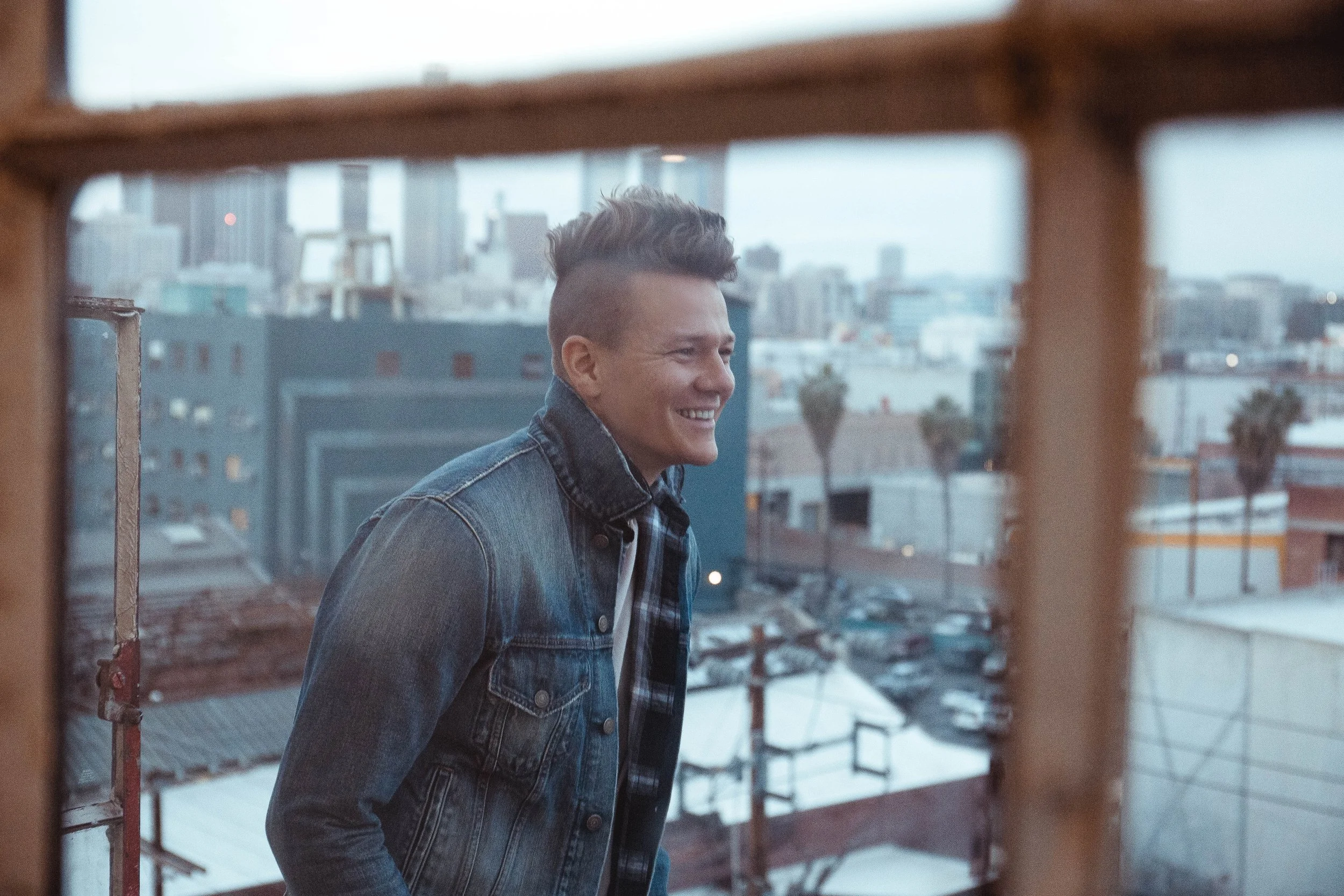 Tyler Ward smiling on a rooftop with a city behind him